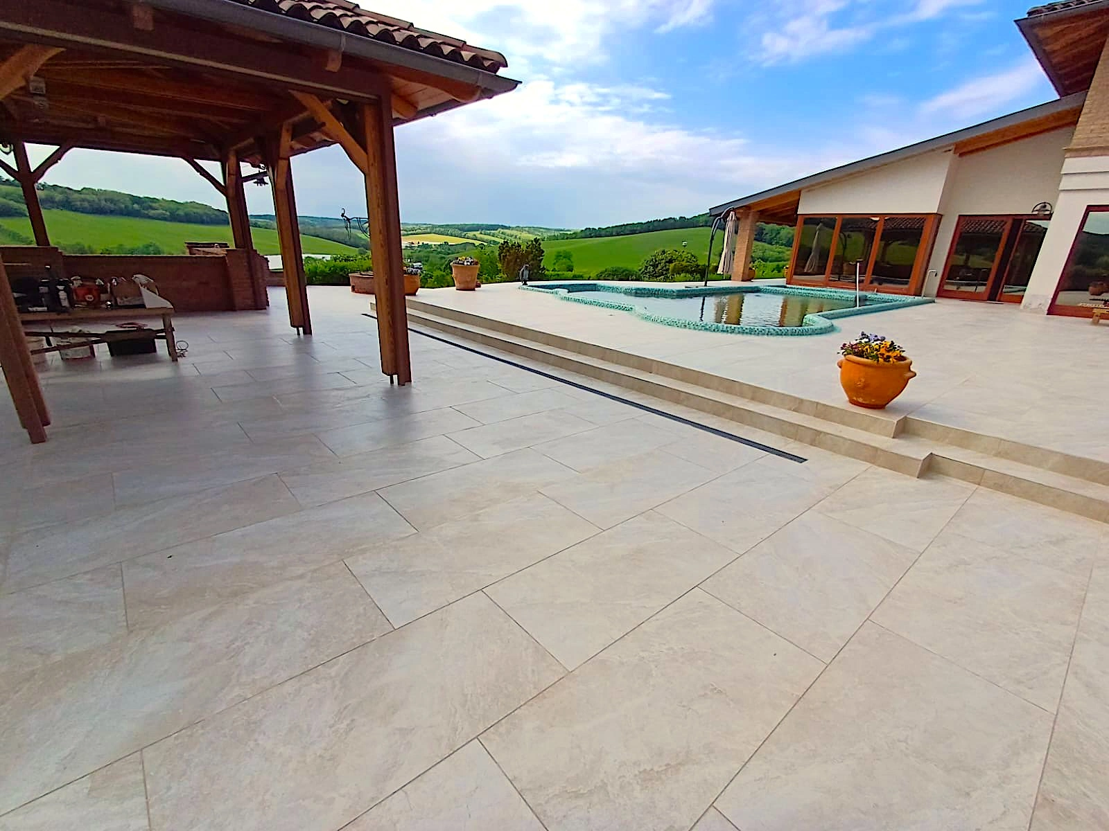 Nagyméretű teraszburkolat medence körül, pergolával és vidéki panorámával.